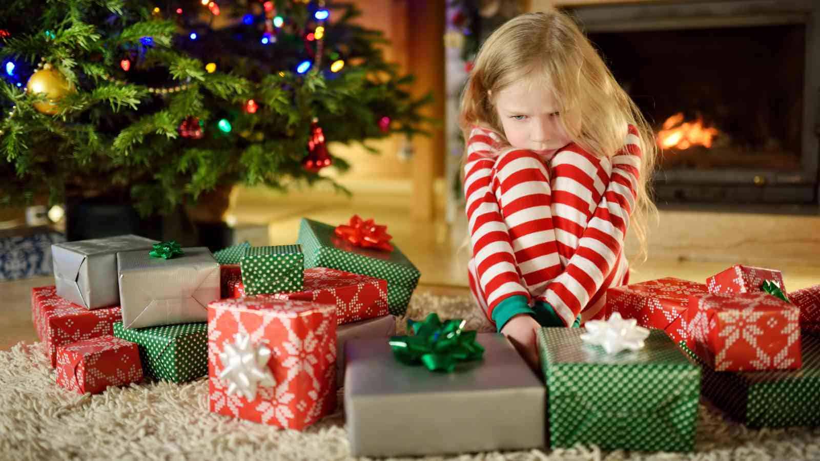 A young girl in striped pajamas sits on the floor, looking sad, surrounded by wrapped gifts near a Christmas tree and fireplace.