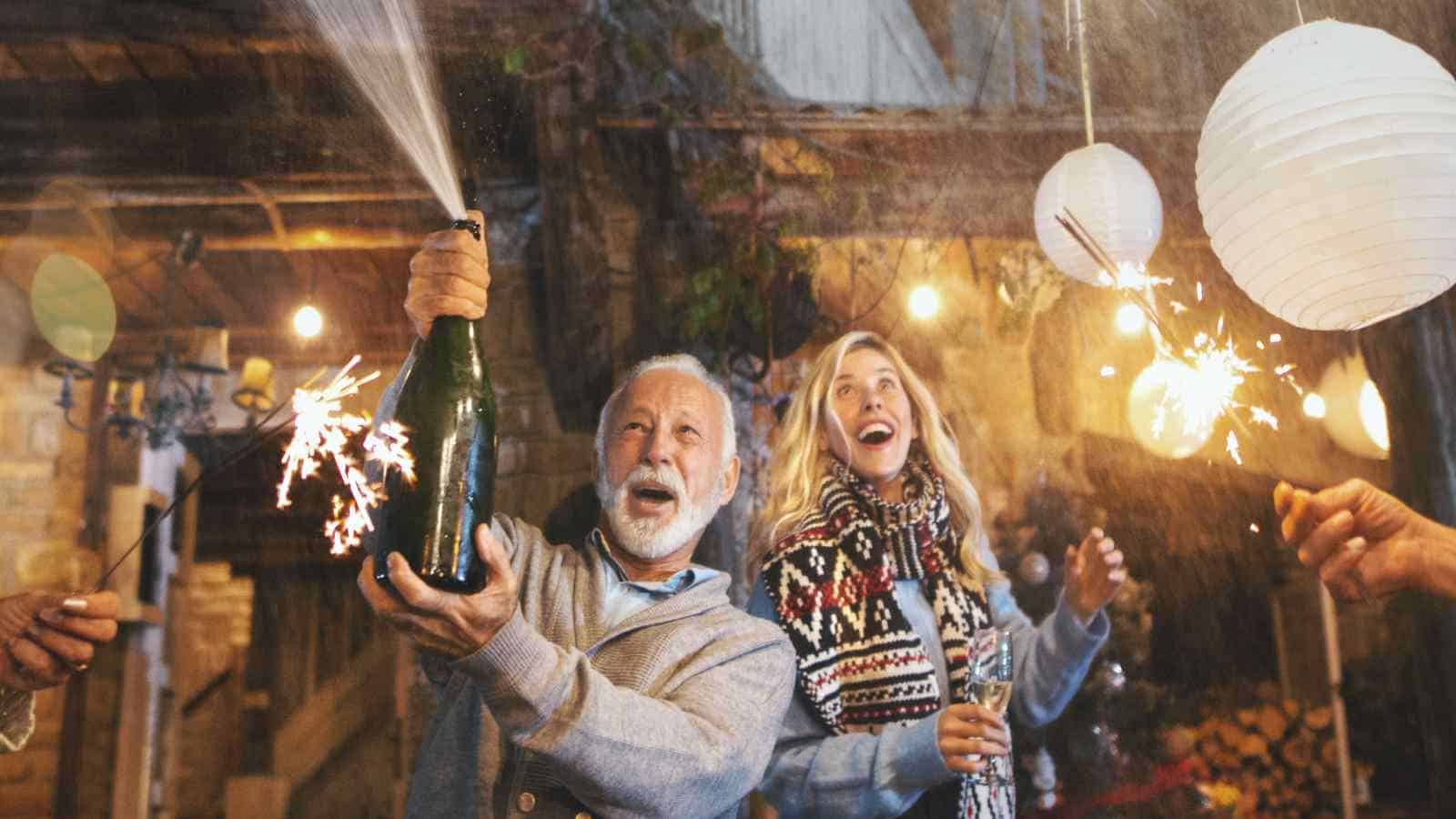An older man pops a champagne bottle while a woman beside him holds a sparkler; they are outdoors at night with string lights and paper lanterns in the background.