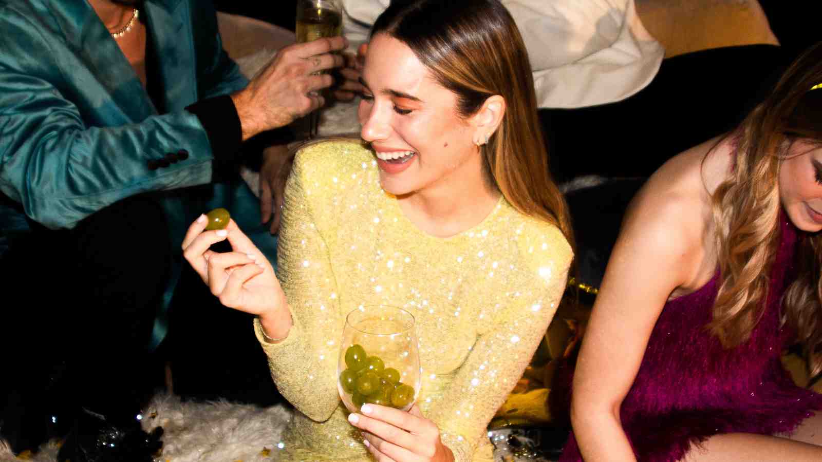 A woman in a yellow sequin dress sits on the floor, smiling and holding green grapes and a glass filled with grapes at a social gathering.