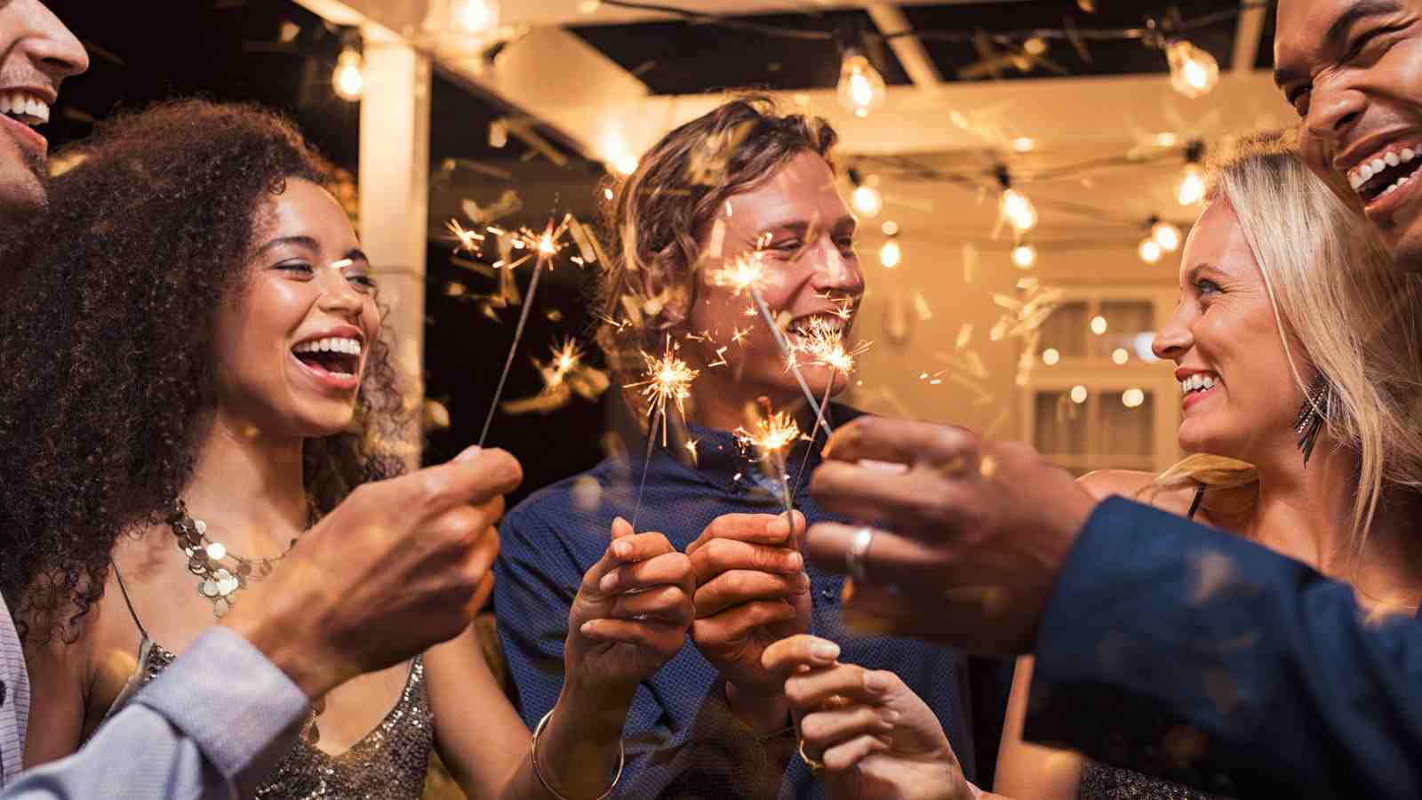 A group of people hold sparklers and smile at each other while standing under string lights at an outdoor gathering.