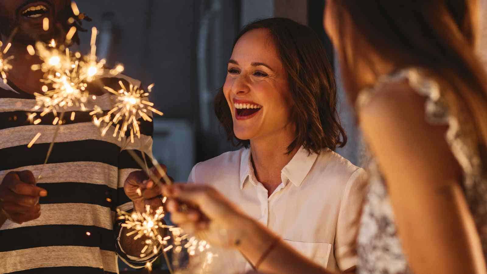 Three people are holding lit sparklers and smiling at night, with one woman in focus laughing and enjoying the moment.