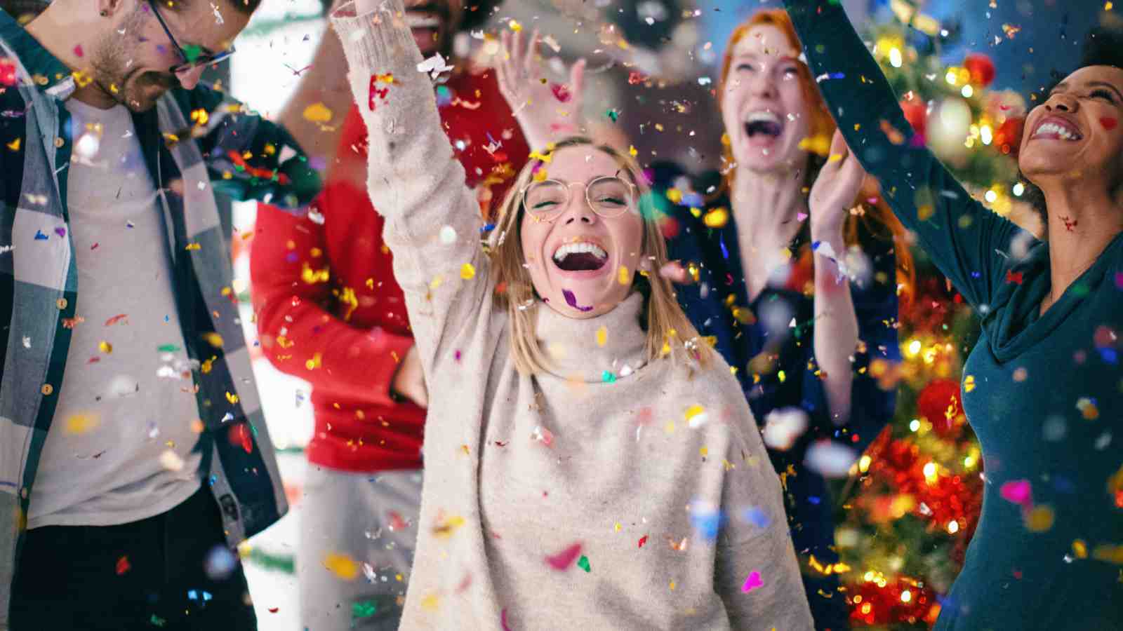A group of people celebrate indoors, smiling and throwing confetti, with festive decorations visible in the background.