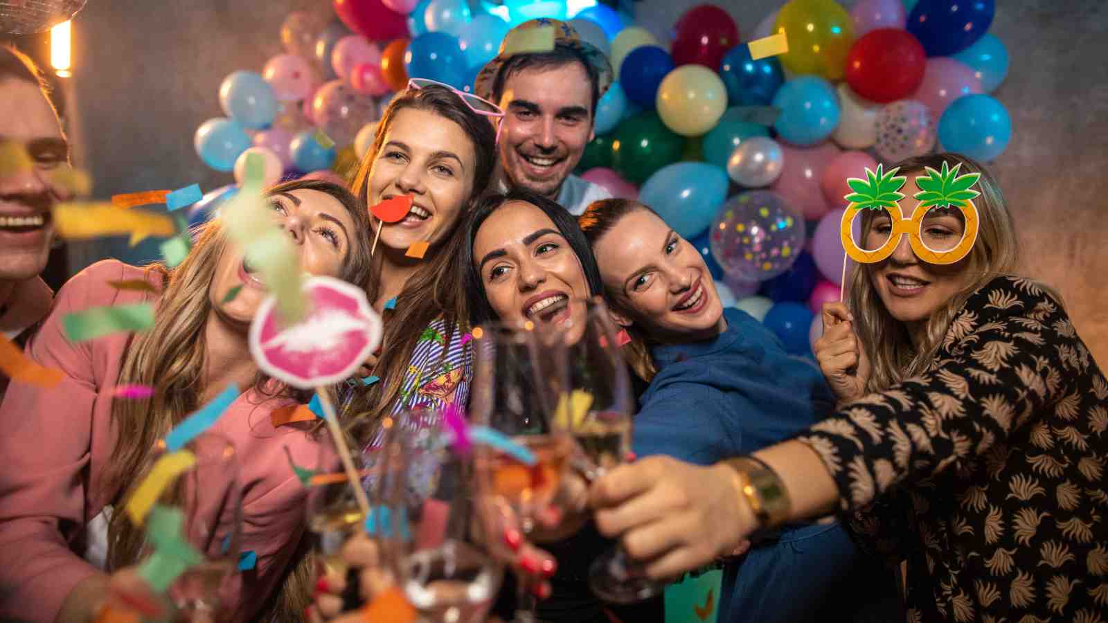 A group of people smiling and posing for a selfie at a party, surrounded by colorful balloons and decorations, some holding props and glasses.