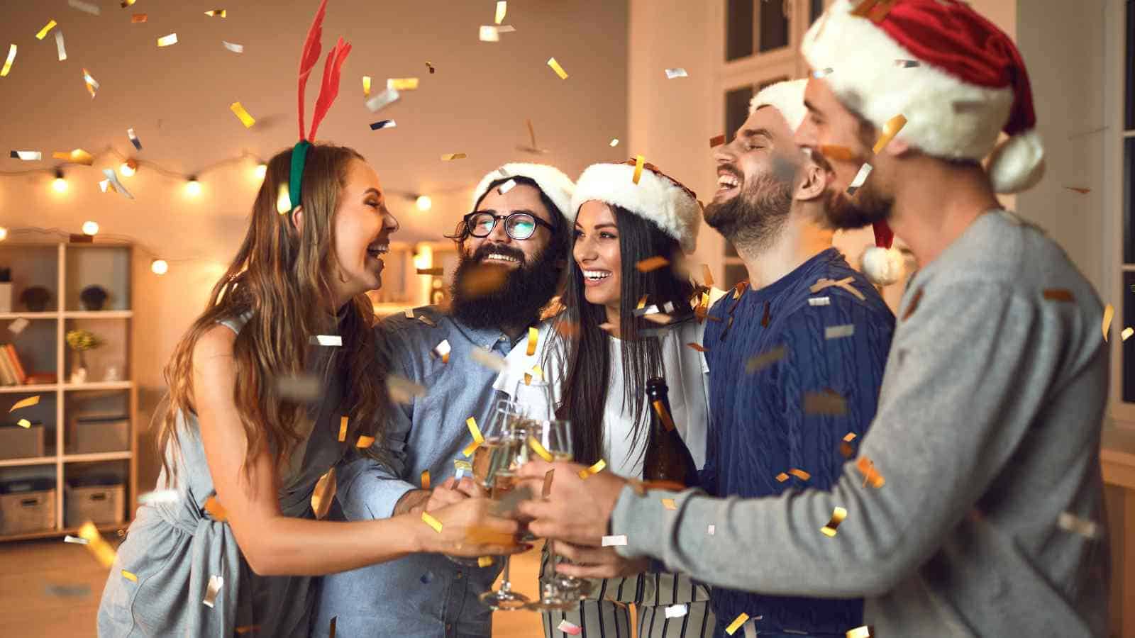 Five people wearing festive hats and holding drinks stand together, smiling and laughing, as gold confetti falls around them in a decorated indoor setting.