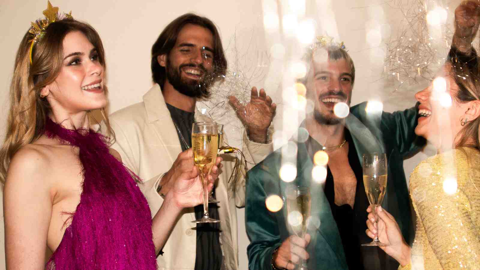 Four people wearing festive outfits and holding champagne glasses celebrate together, smiling and laughing, with sparkling decorations in the foreground.