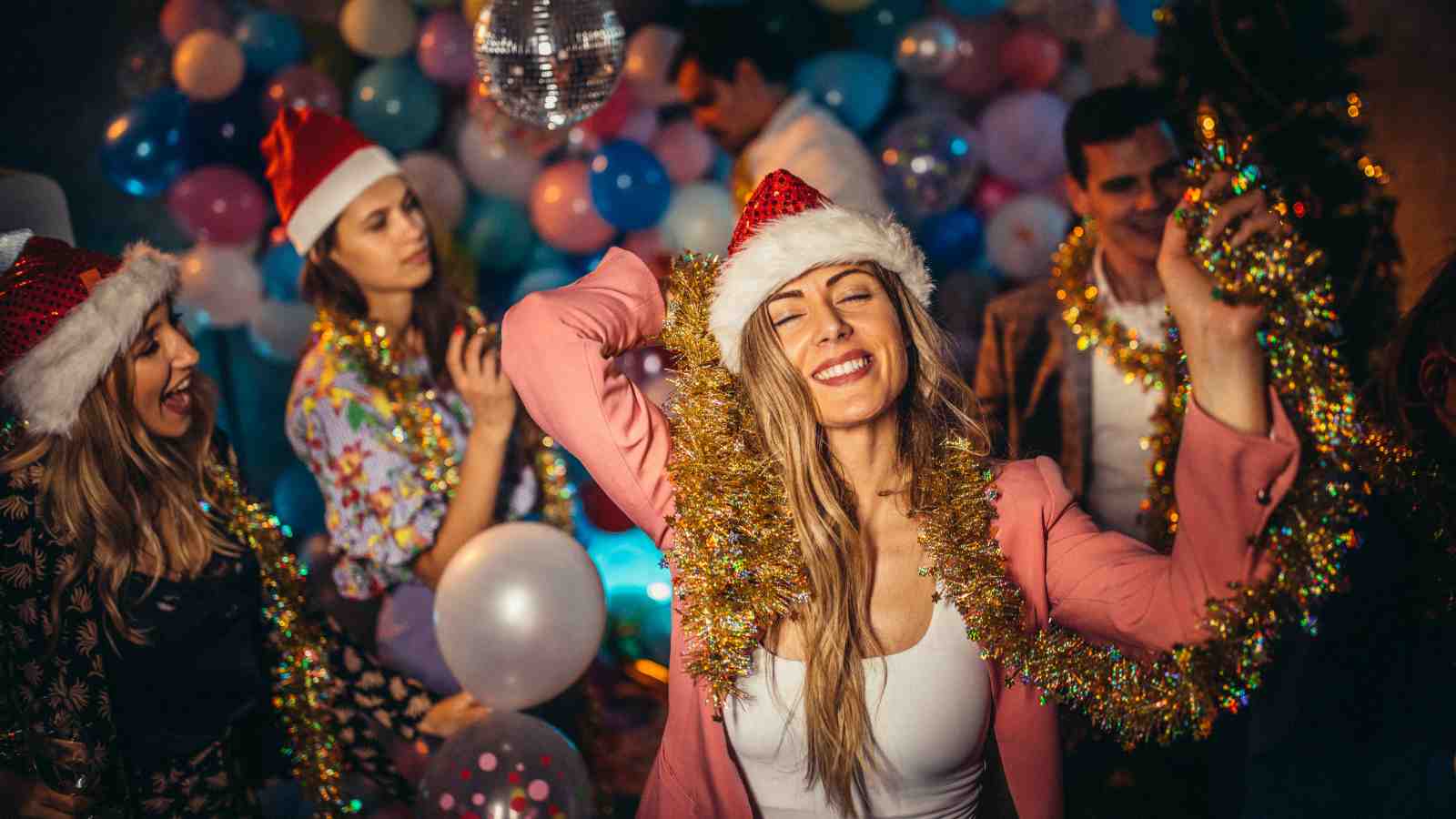 People wearing Santa hats and festive garlands dance and celebrate at a party decorated with balloons and a disco ball.