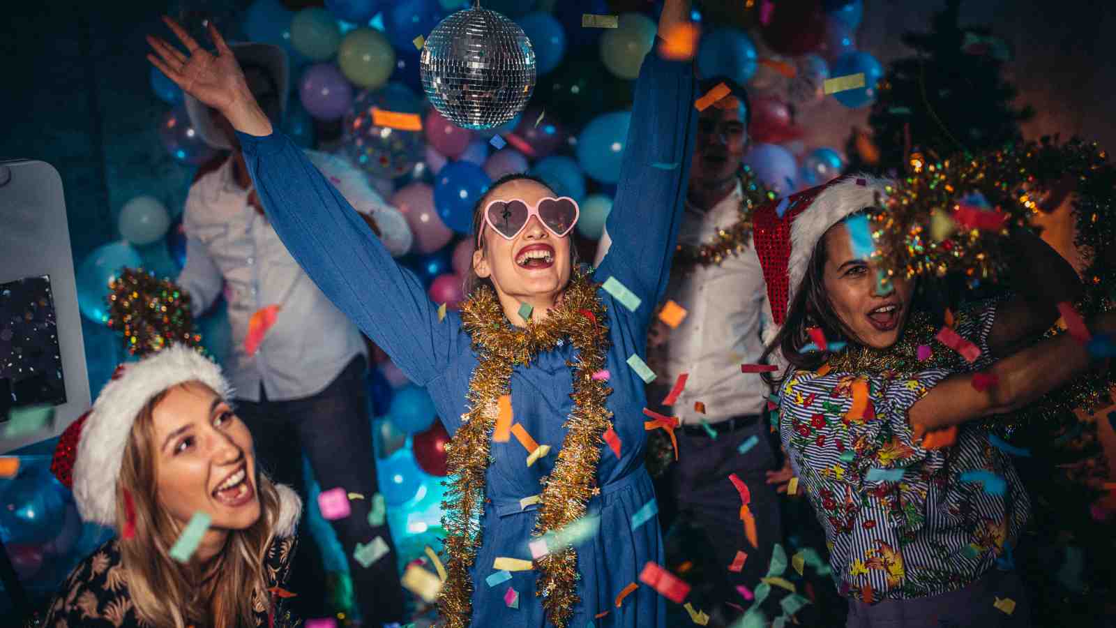 Three people wearing festive hats and tinsel celebrate at a party, surrounded by balloons, confetti, and a disco ball, with more people in the background.