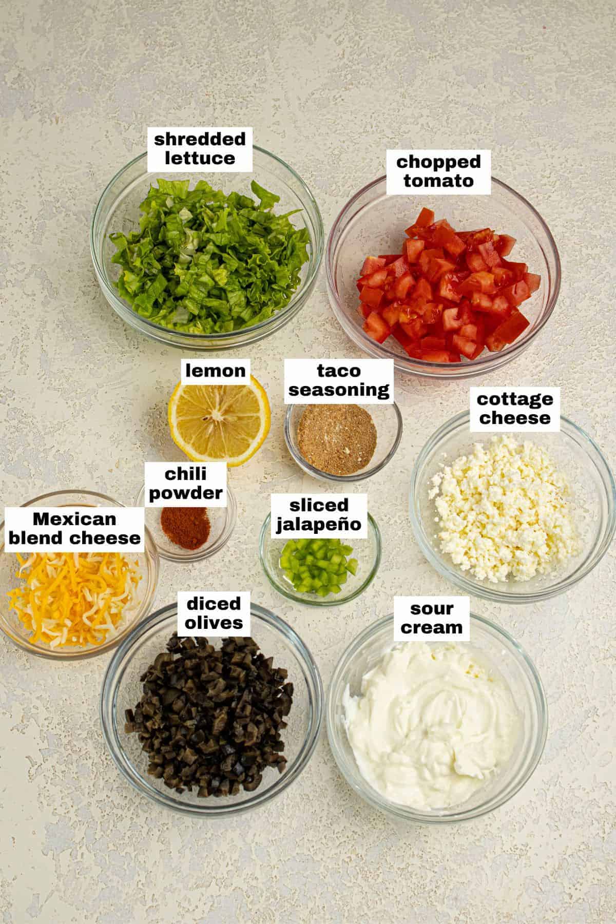 Ten glass bowls containing shredded lettuce, chopped tomato, lemon, taco seasoning, cottage cheese, Mexican blend cheese, chili powder, sliced jalapeño, diced olives, and sour cream.