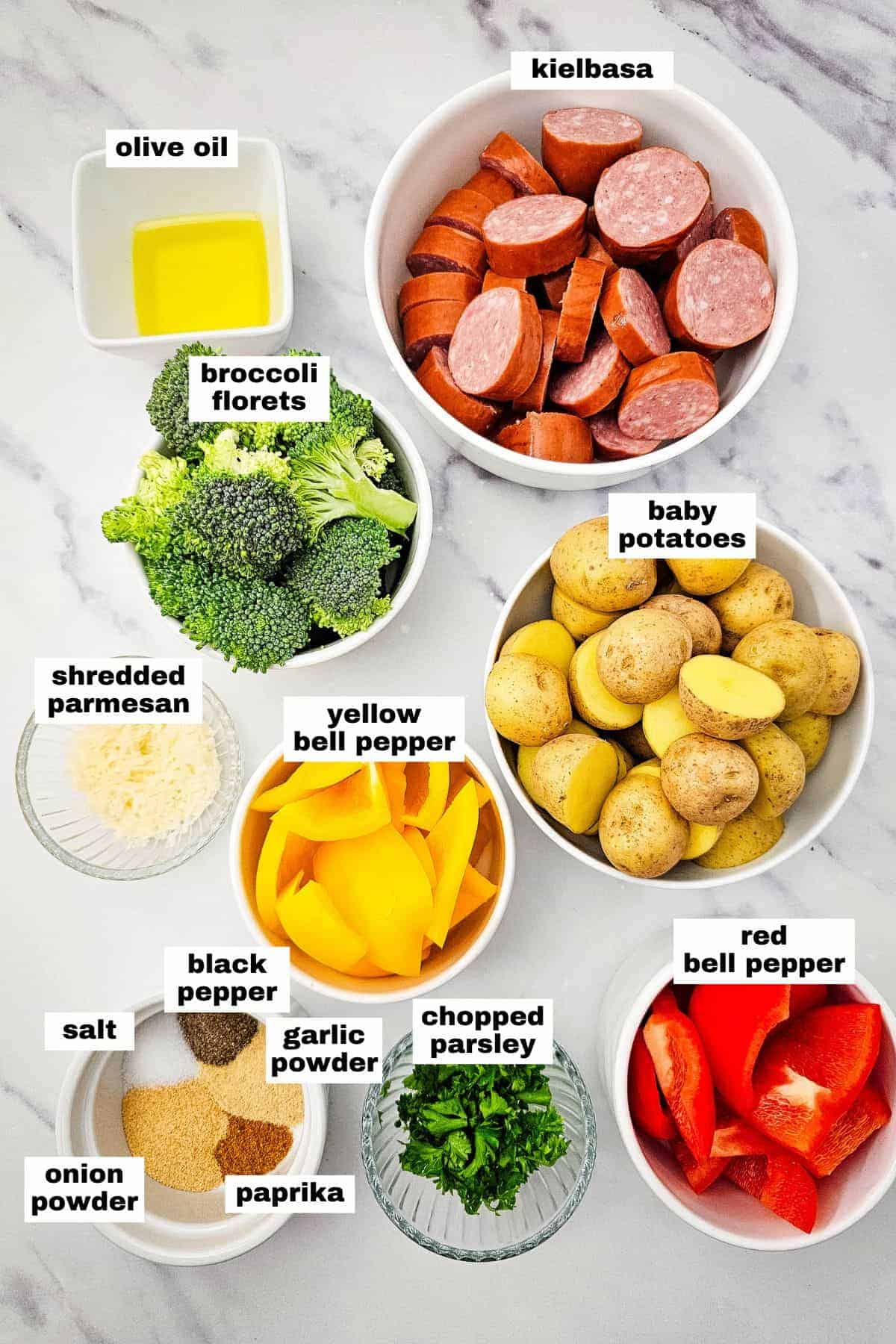 Bowls on a countertop contain labeled ingredients: olive oil, kielbasa, broccoli florets, baby potatoes, yellow and red bell peppers, shredded parmesan, parsley, and seasonings.