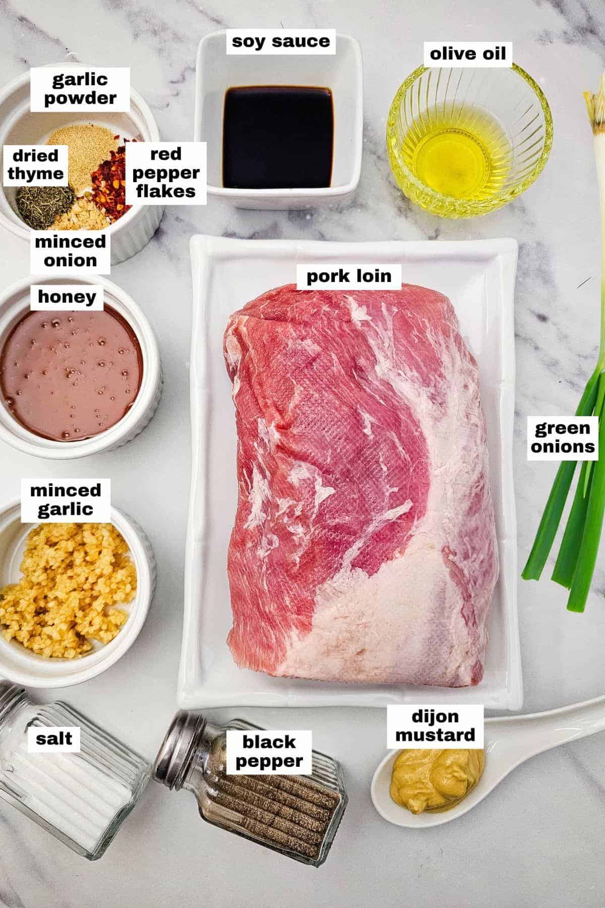 Raw pork loin surrounded by labeled ingredients including garlic powder, dried thyme, red pepper flakes, minced onion, honey, salt, black pepper, soy sauce, olive oil, Dijon mustard, and green onions.