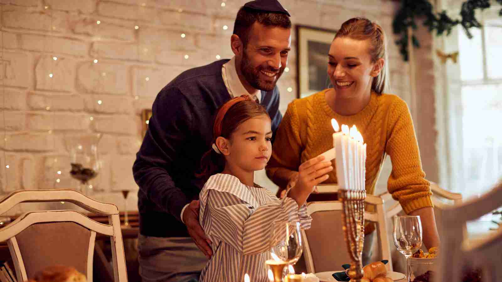 A young girl lights candles on a menorah, with an adult man and woman smiling beside her at a decorated dining table.