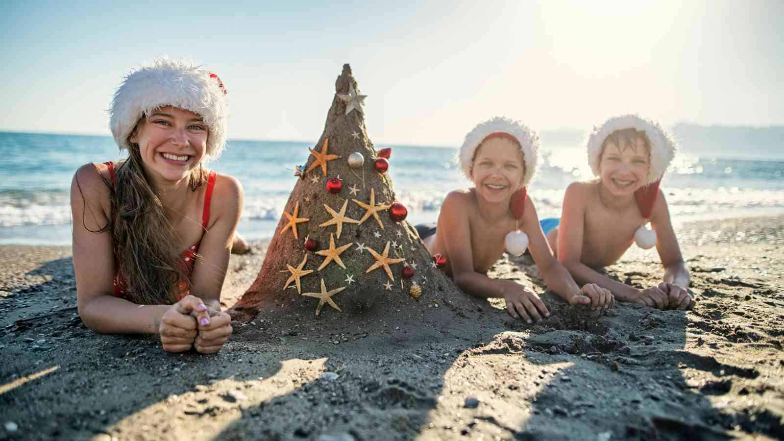 Three children in Santa hats lie on a beach next to a sand Christmas tree decorated with starfish and ornaments, with the sea in the background.