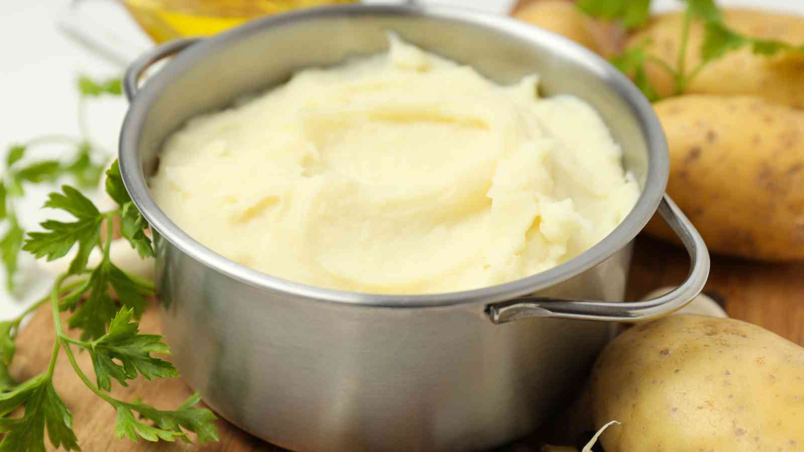 A metal pot filled with creamy mashed potatoes, surrounded by whole potatoes and fresh parsley on a wooden surface.
