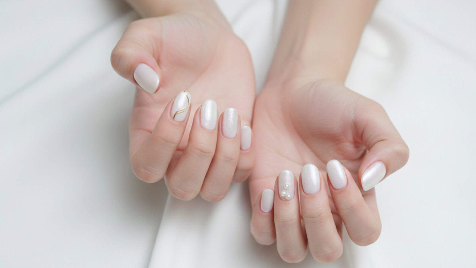Two hands showing manicured nails painted in pale pink with accents; one nail features a gold line design and another has small pearl decorations.
