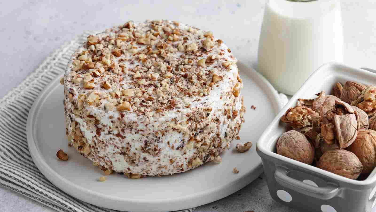 A round cake covered in white frosting and chopped walnuts sits on a plate beside a bowl of whole walnuts and a glass of milk.