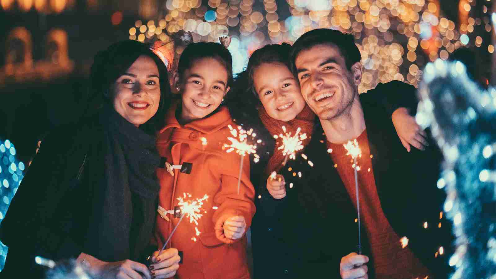 A family of four holds sparklers and smiles at the camera, with festive lights and a bokeh background.