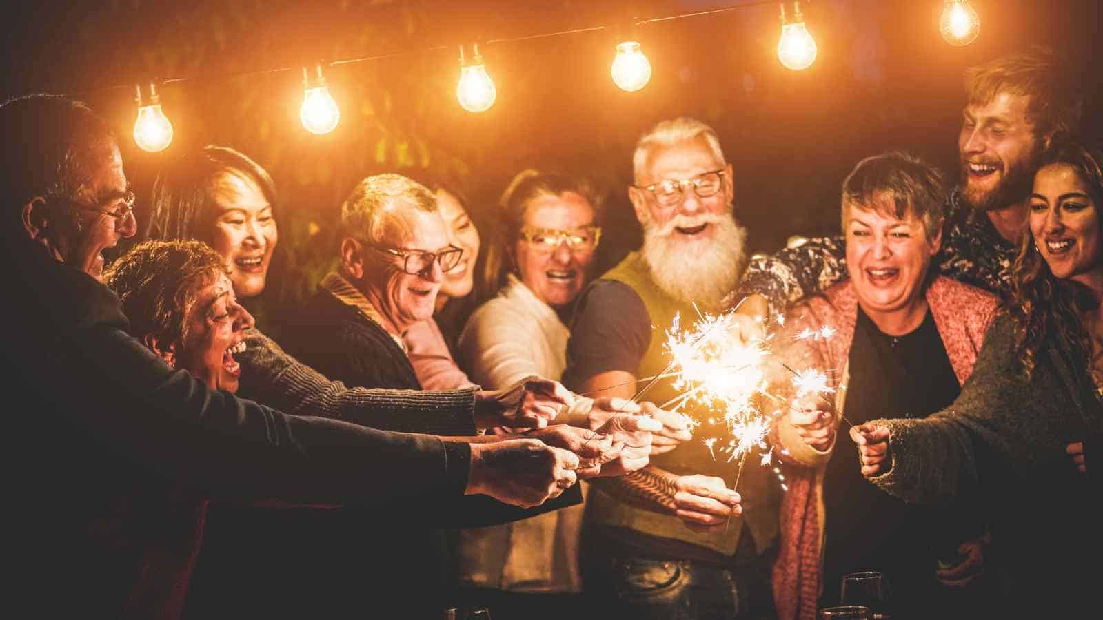 A group of people stand close together at night, holding sparklers and smiling under a string of hanging lights.