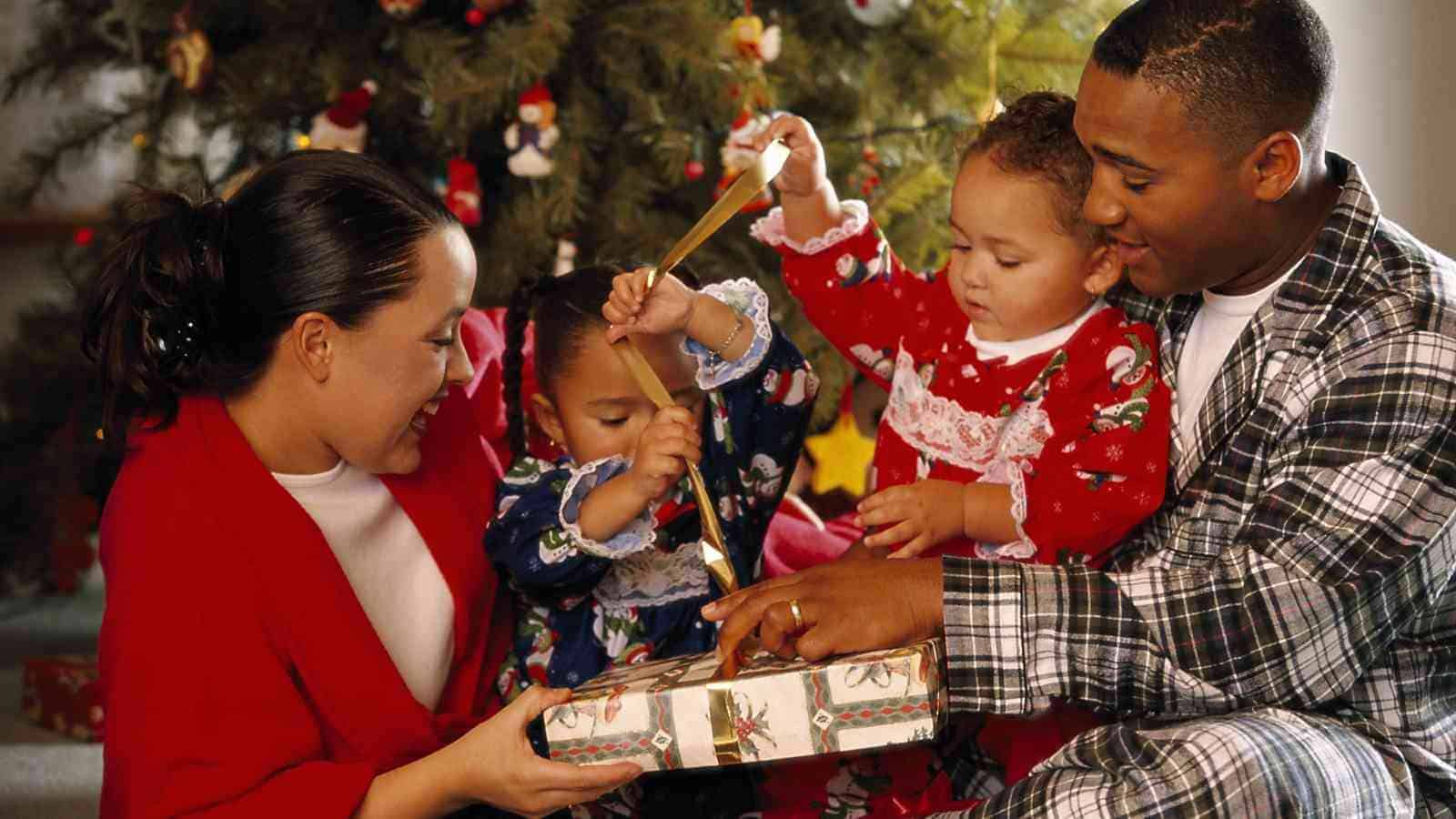 A family of four sits by a decorated Christmas tree, unwrapping a present together. The children and adults are dressed in festive pajamas.