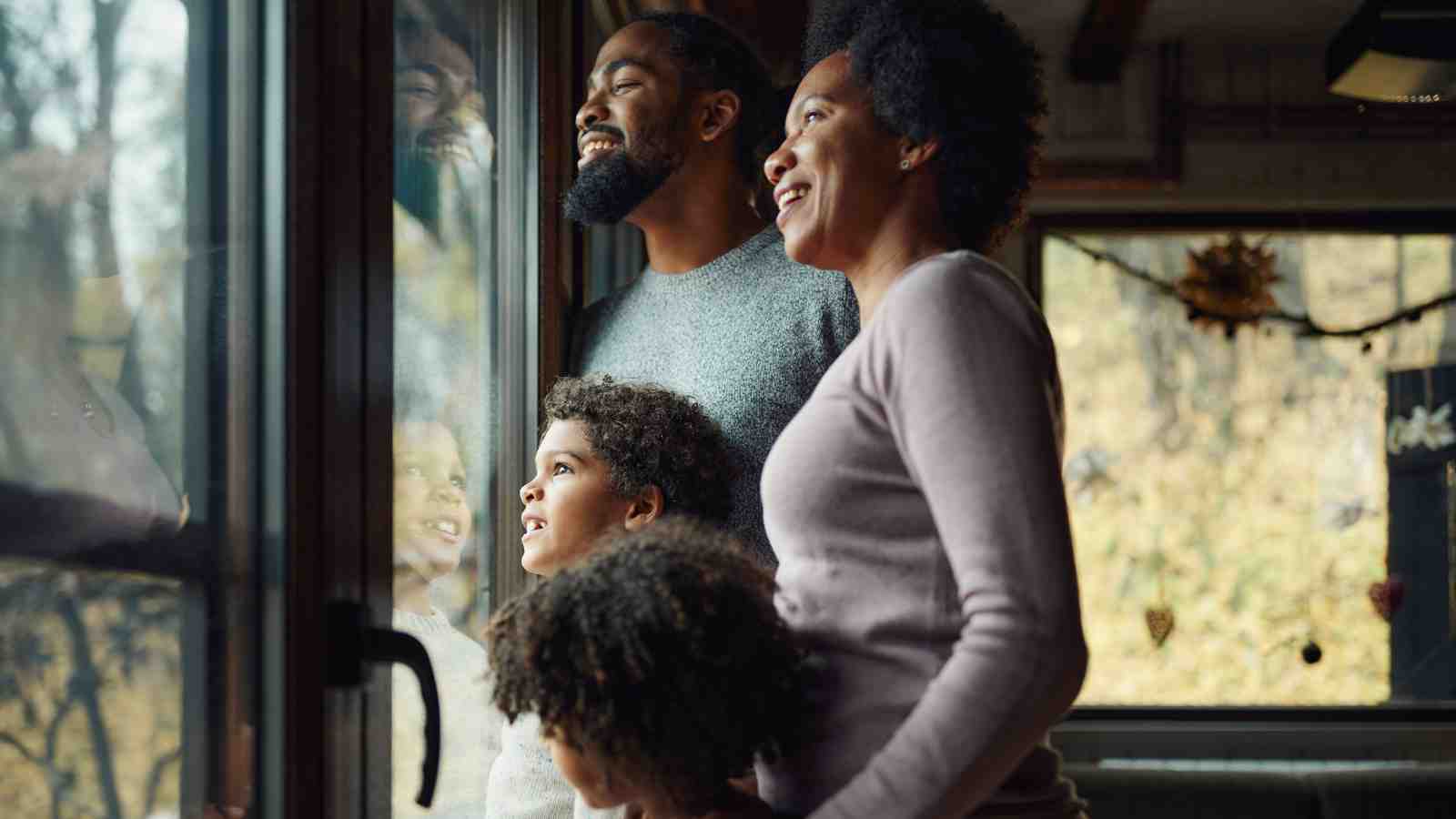 A family of four stands indoors near a window, looking outside and smiling. Natural light comes through the glass, with trees visible outside.