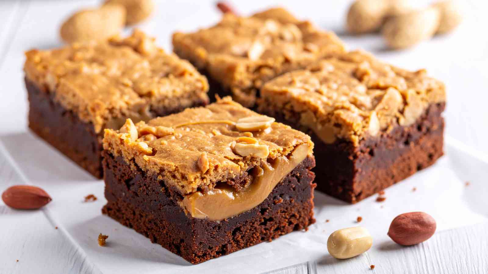Three square pieces of chocolate and peanut butter brownies with a layer of peanuts and peanut butter on top, placed on parchment paper with whole peanuts around.
