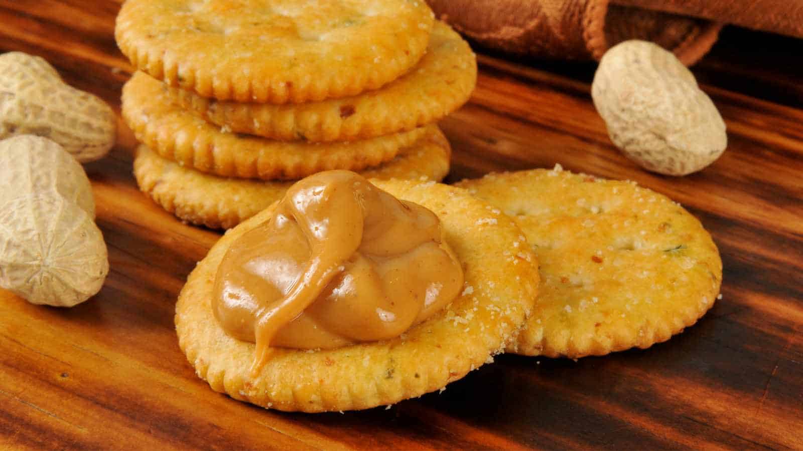 Round crackers on a wooden surface, with one cracker topped with a dollop of creamy peanut butter. Whole peanuts are also visible nearby.