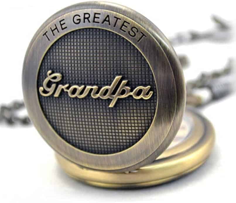 A brass pocket watch with "Grandpa" in script and "The Greatest" engraved above it, shown closed with part of the chain visible in the background.