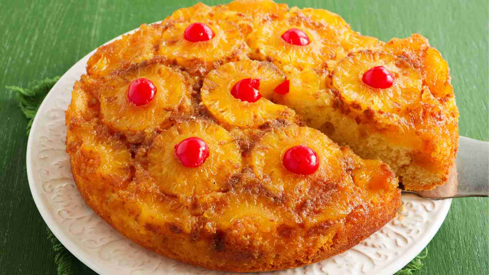 A pineapple upside-down cake topped with pineapple rings and maraschino cherries, with one slice being served.