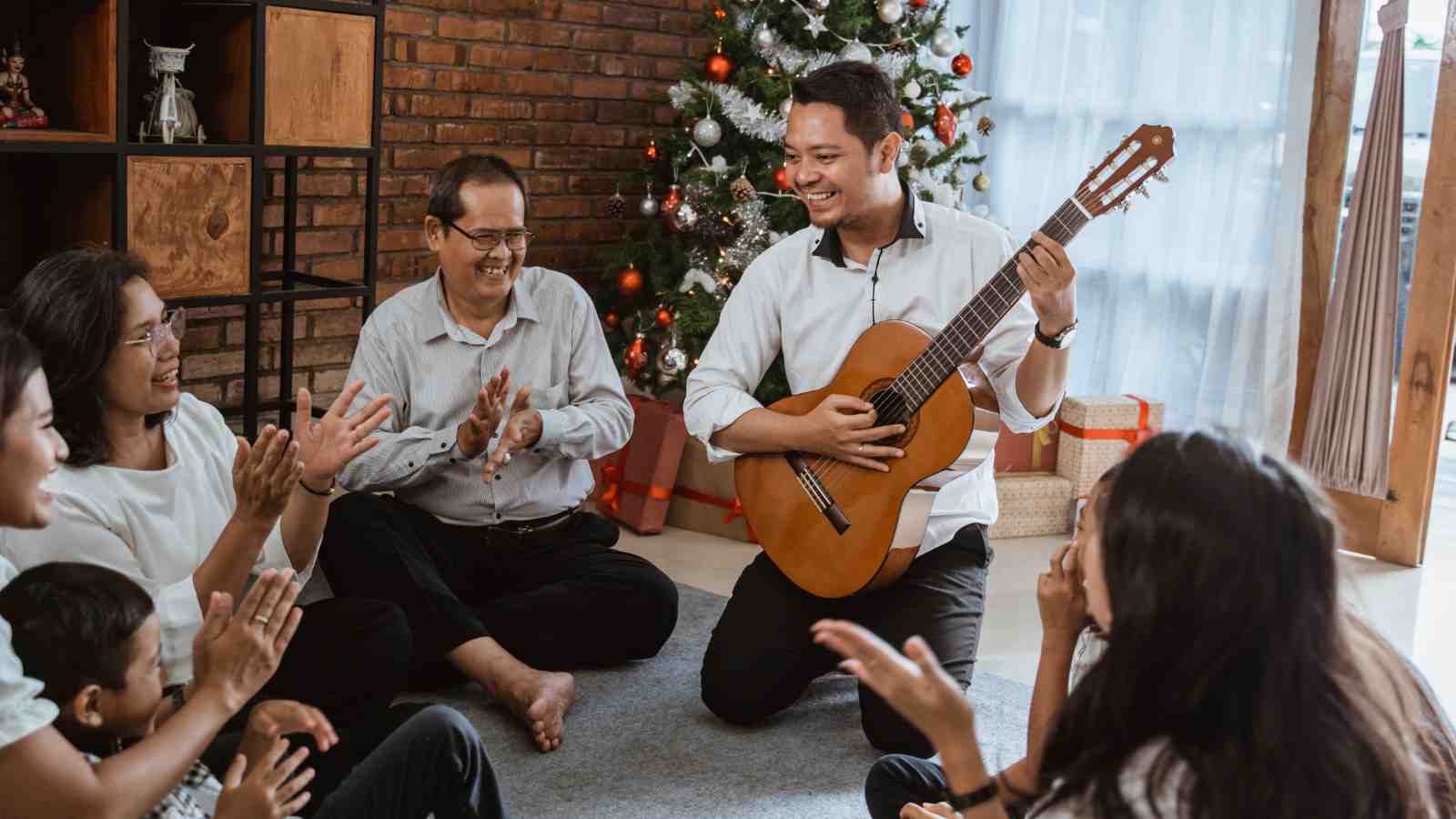A group of people sit on the floor near a decorated Christmas tree, clapping while a man plays a guitar and smiles.