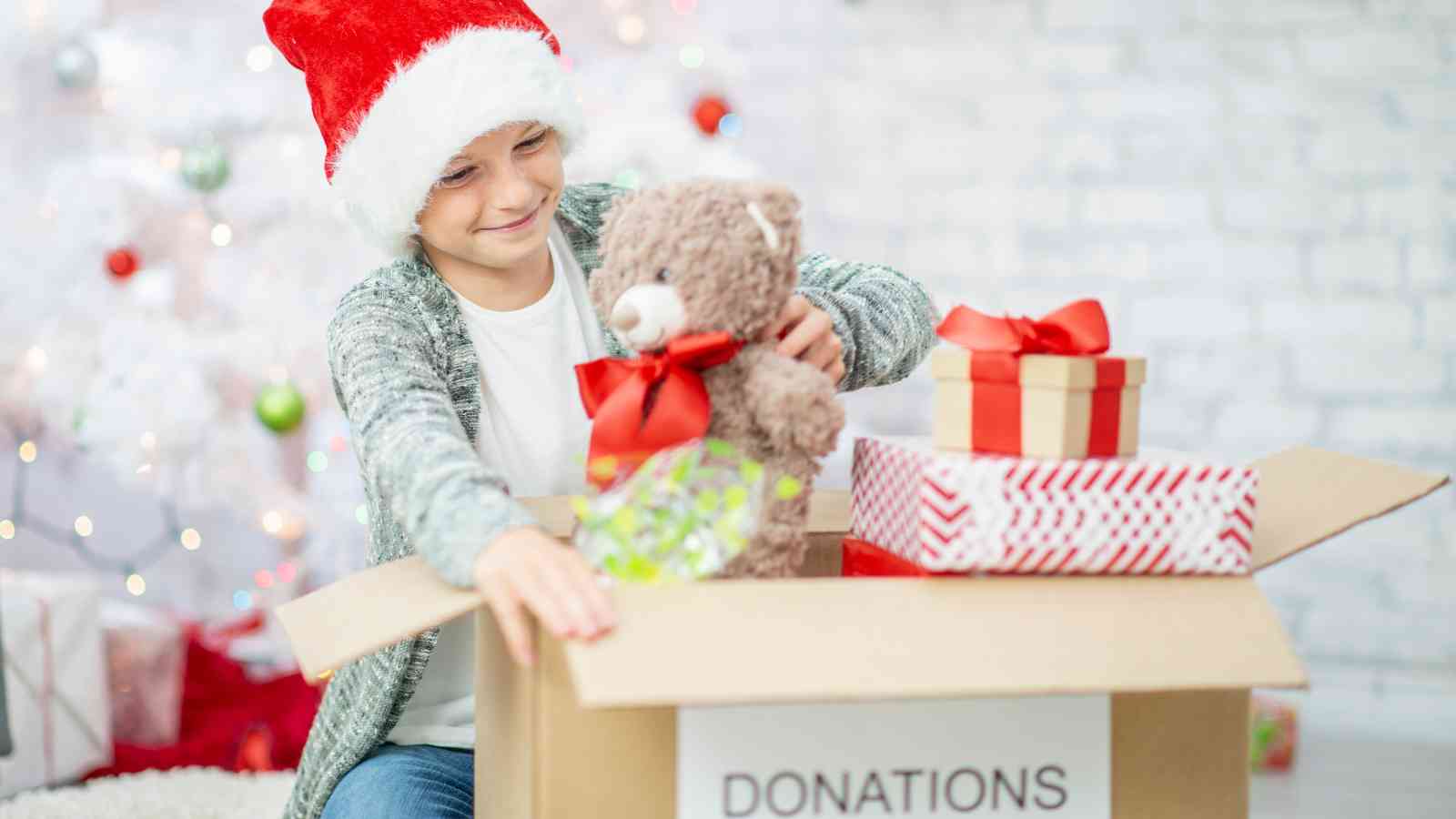 A child wearing a Santa hat places a teddy bear and wrapped gifts into a cardboard box labeled "Donations" in front of a decorated Christmas tree.