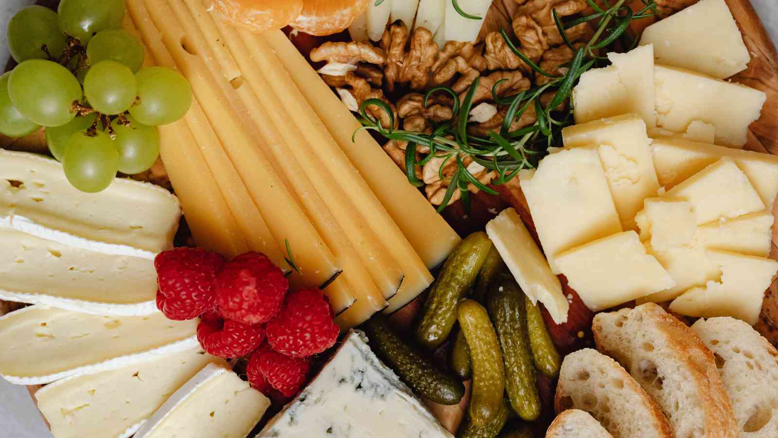 A cheese board with assorted cheeses, sliced bread, grapes, raspberries, walnuts, pickles, and sprigs of rosemary arranged on a wooden surface.
