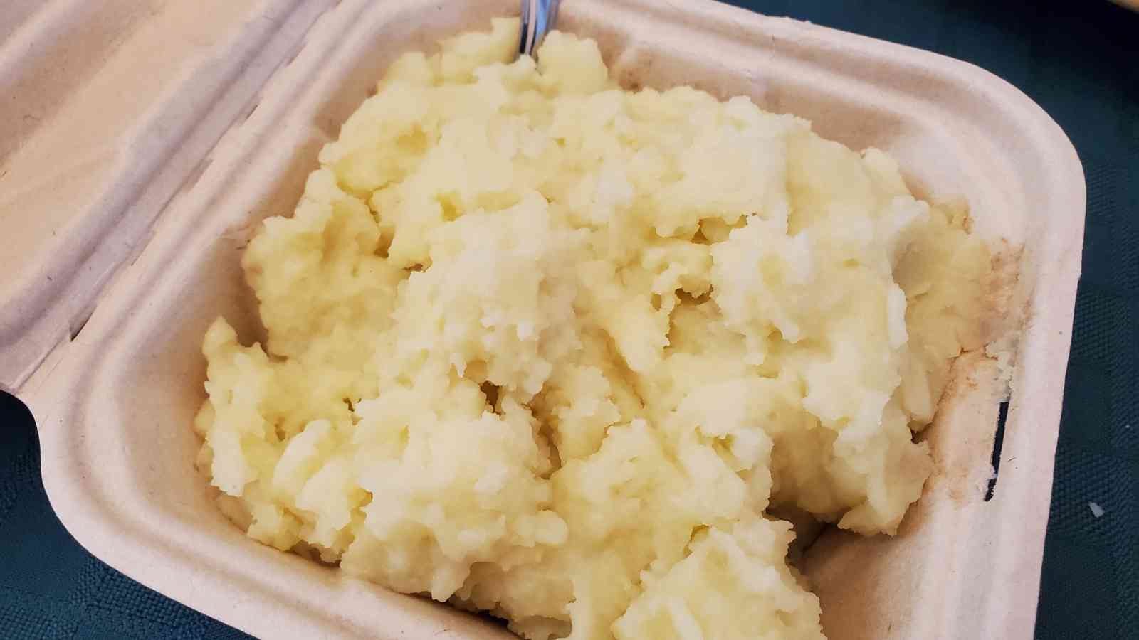 A takeout container filled with mashed potatoes, with part of a metal utensil visible at the top.