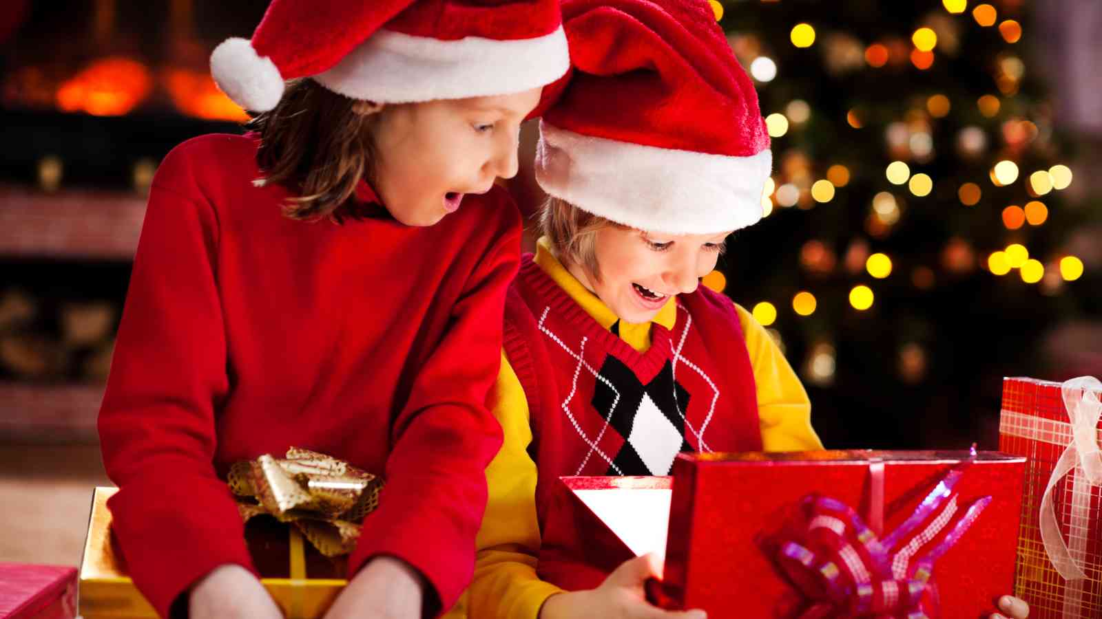 Two children in Santa hats and festive clothes excitedly open Christmas presents, with a decorated tree and lights in the background.