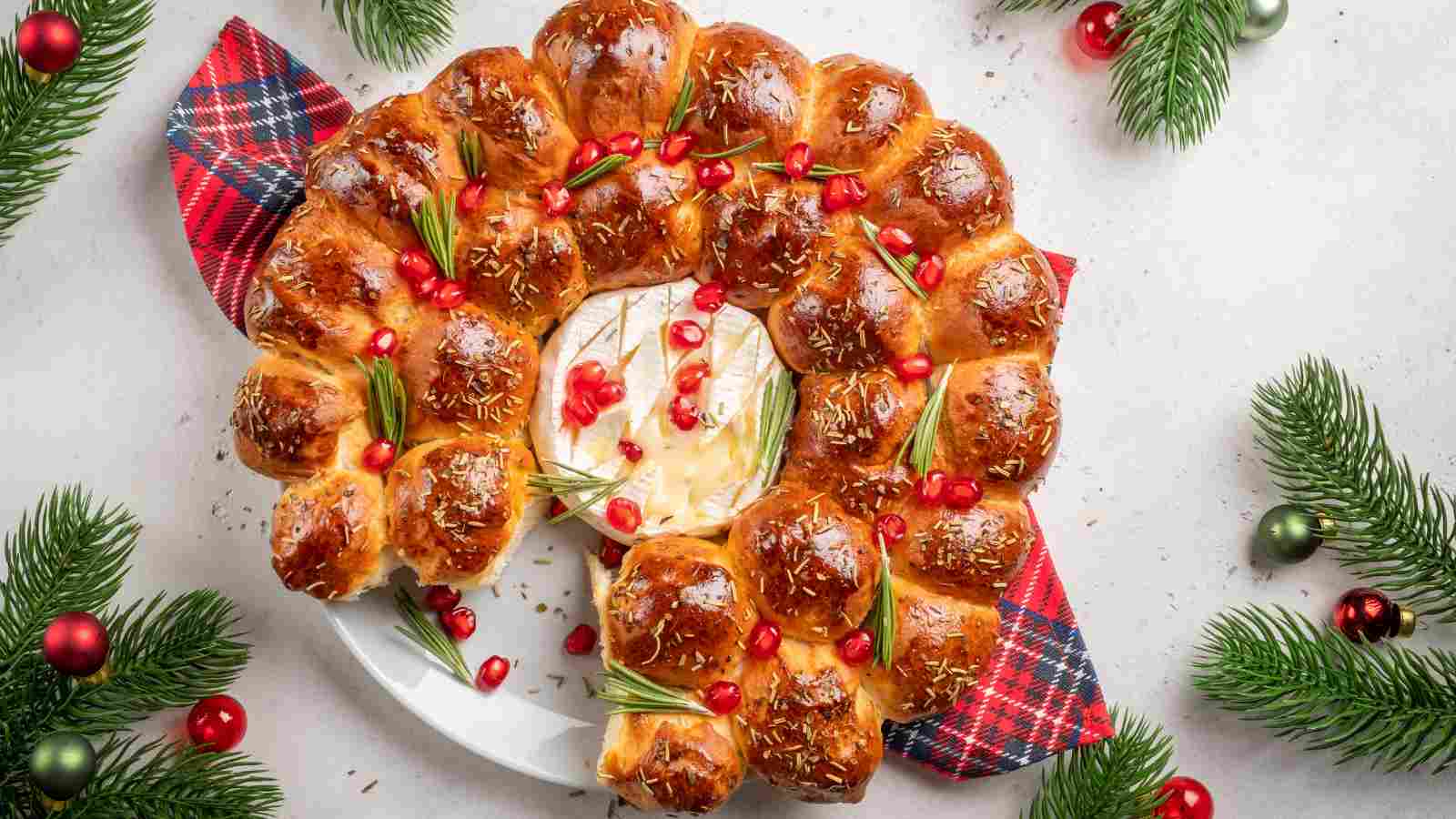 A round pull-apart bread wreath with rosemary, pomegranate seeds, and baked brie in the center, garnished with pine branches and ornaments.