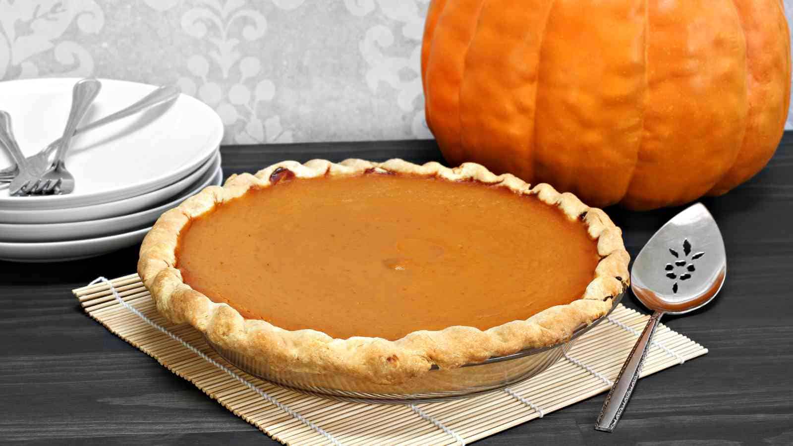 A whole pumpkin pie in a glass dish sits on a bamboo mat next to a large pumpkin, plates with forks, and a pie server on a dark table.