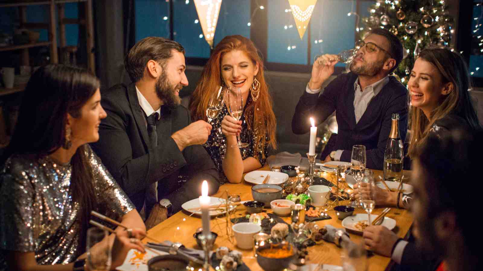 Six people dressed in festive attire sit around a dinner table, eating, drinking, and smiling together in a warmly lit room decorated with lights.