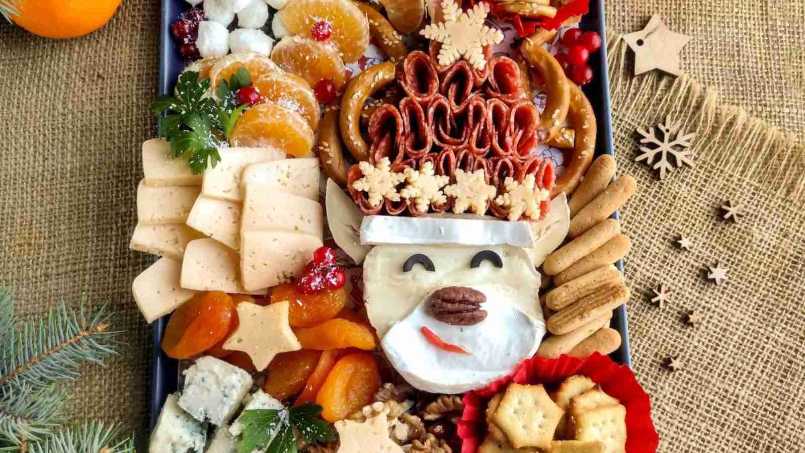 A festive charcuterie board features cheese, crackers, dried fruit, pretzels, and orange slices, arranged with brie cheese shaped like a bear’s face, decorated with nuts and herbs. Oranges and pine branches are nearby.