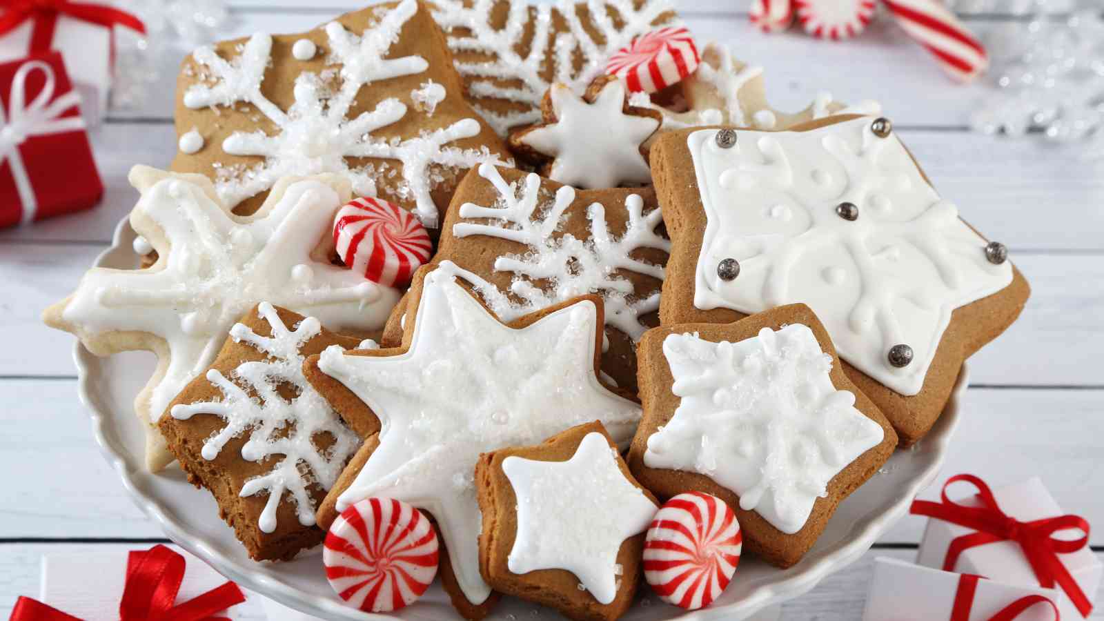 A plate of snowflake-shaped gingerbread cookies with white icing, surrounded by red and white peppermint candies on a white table.
