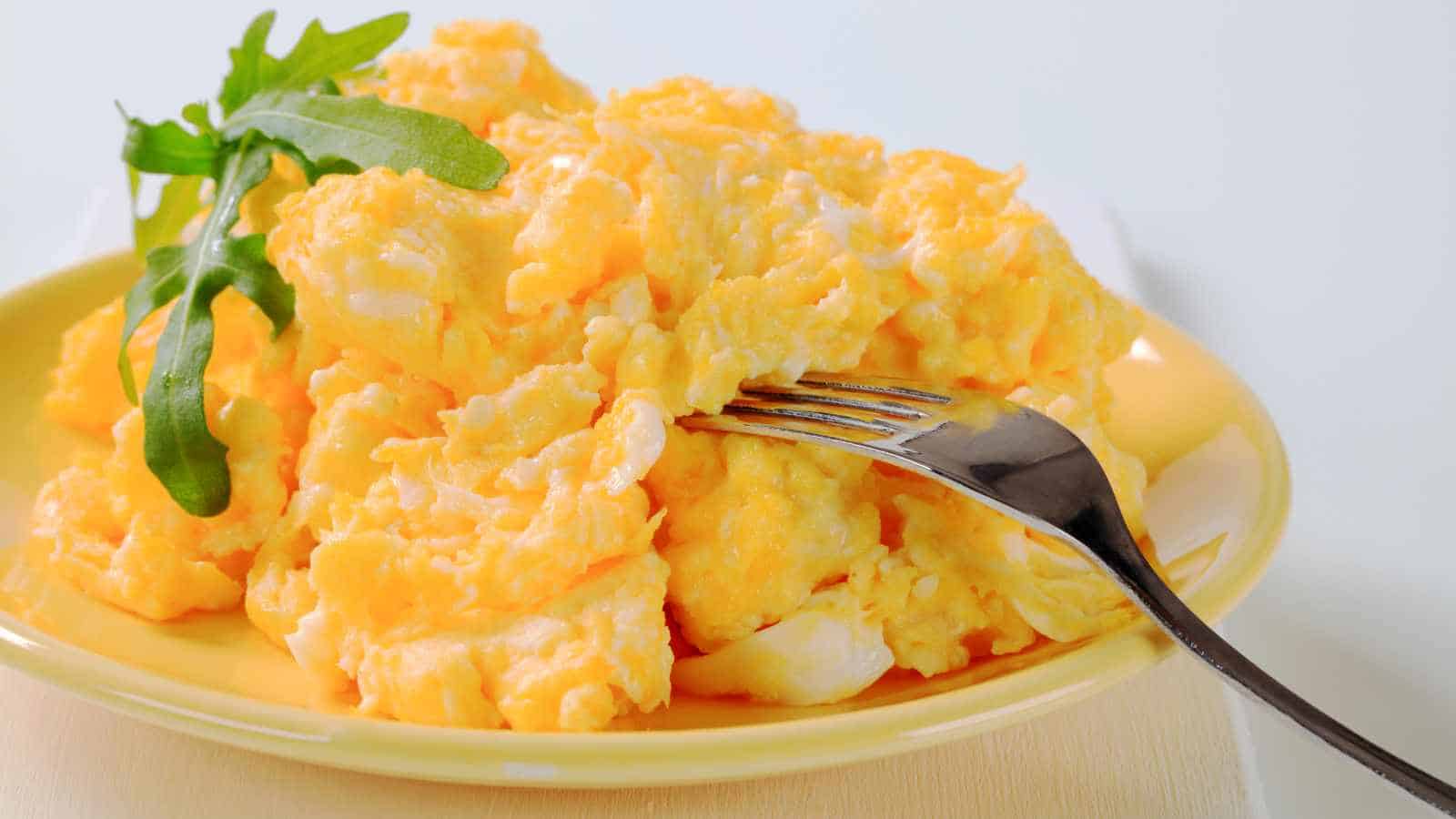 A plate of scrambled eggs garnished with a few arugula leaves, with a fork resting on the plate.