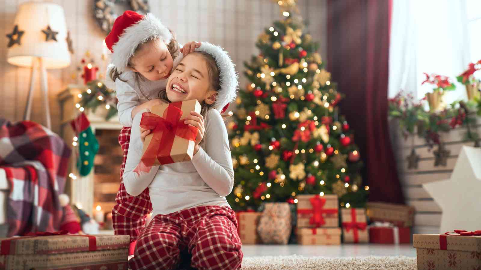 Two children in Santa hats and plaid pajamas exchange gifts and hug in front of a decorated Christmas tree surrounded by presents in a cozy living room.