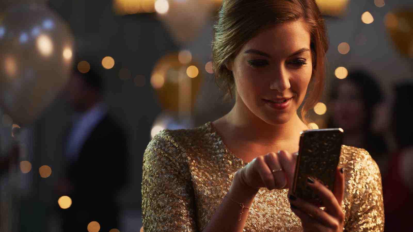 A woman in a gold dress uses her smartphone at a party, with balloons and blurred lights in the background.