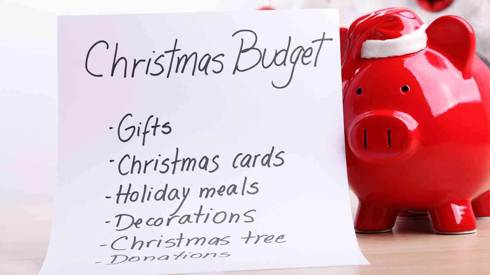 A handwritten Christmas budget list sits next to a red piggy bank wearing a Santa hat. The list includes gifts, cards, meals, decorations, tree, and donations.