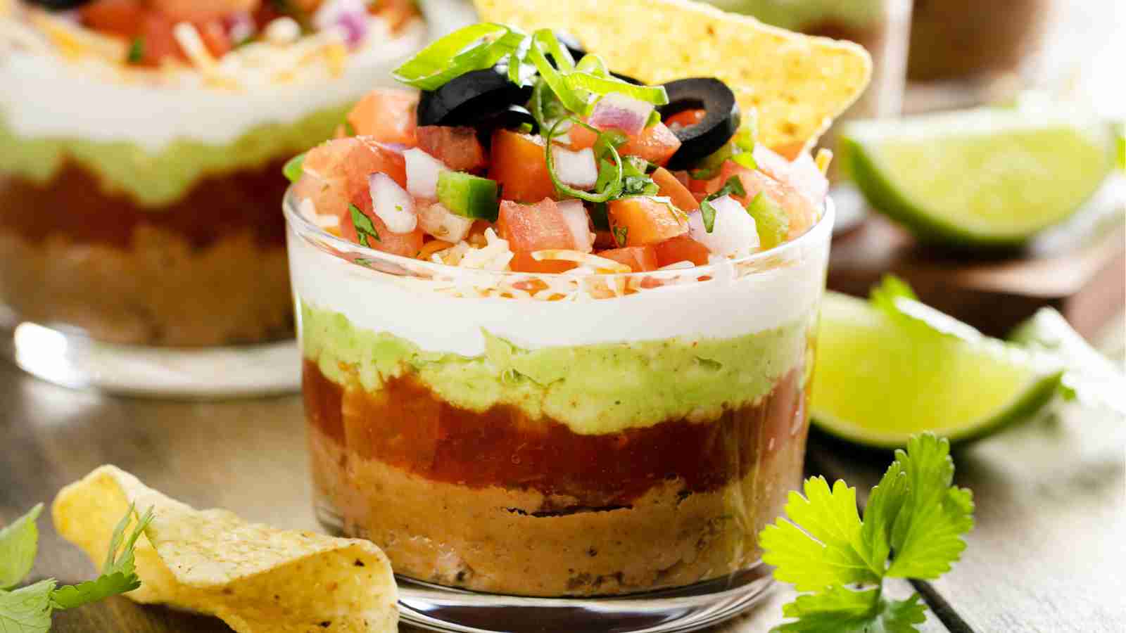 A glass cup layered with refried beans, salsa, guacamole, sour cream, shredded cheese, diced tomatoes, black olives, and green onions, garnished with tortilla chips.