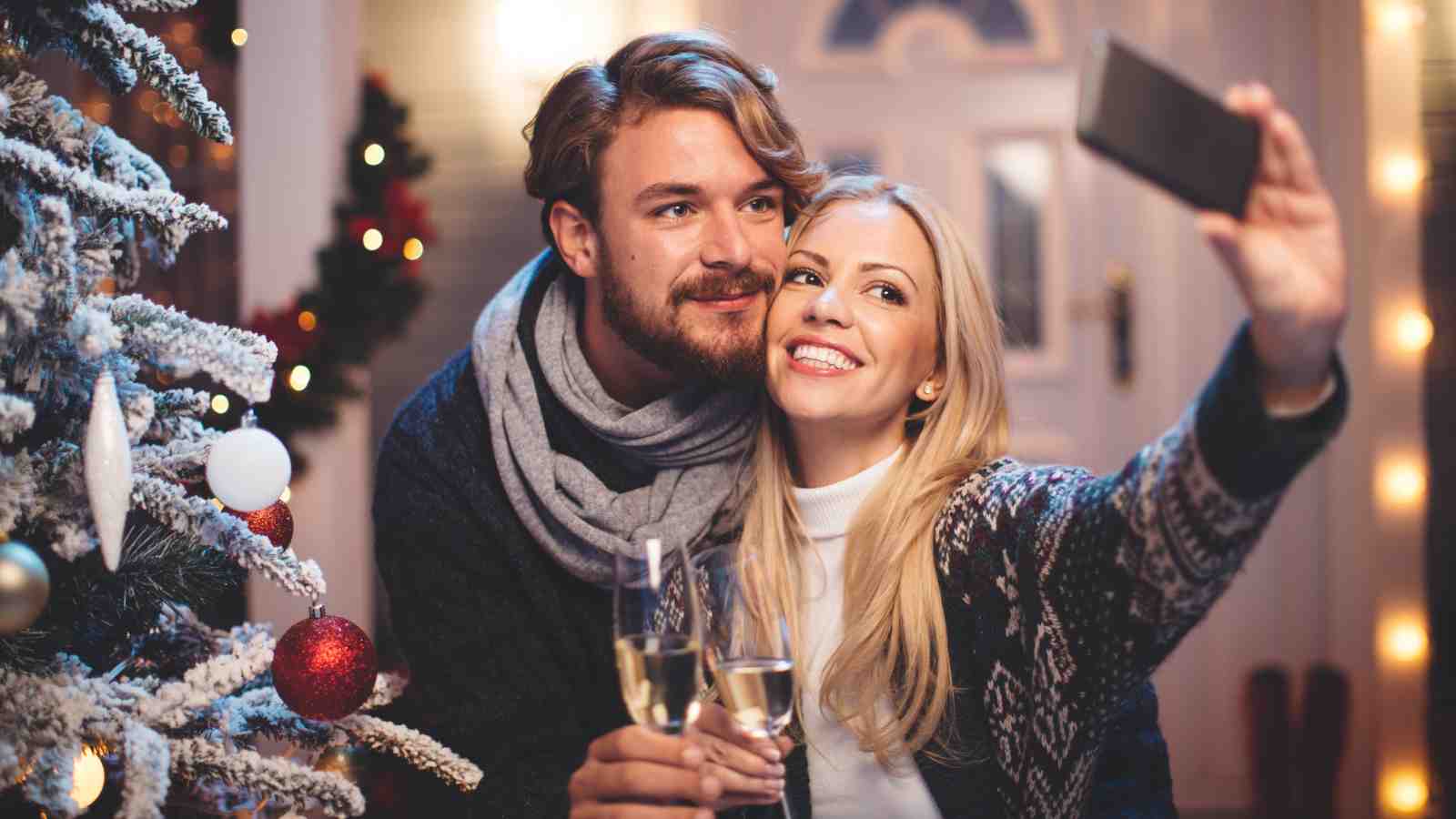 A couple takes a selfie together by a decorated Christmas tree, holding glasses of white wine and smiling.