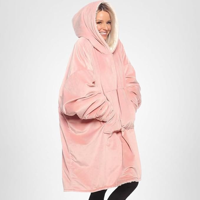 A person wearing an oversized, light pink hooded blanket sweatshirt with hands in the front pocket, standing against a plain light background.