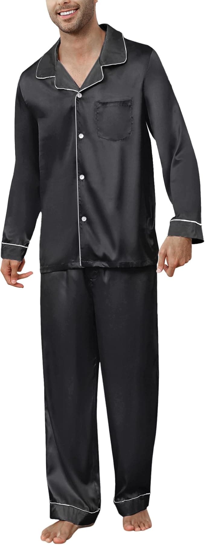 A man wearing a black satin pajama set with white piping, consisting of a long-sleeve button-up top and matching pants, standing barefoot.