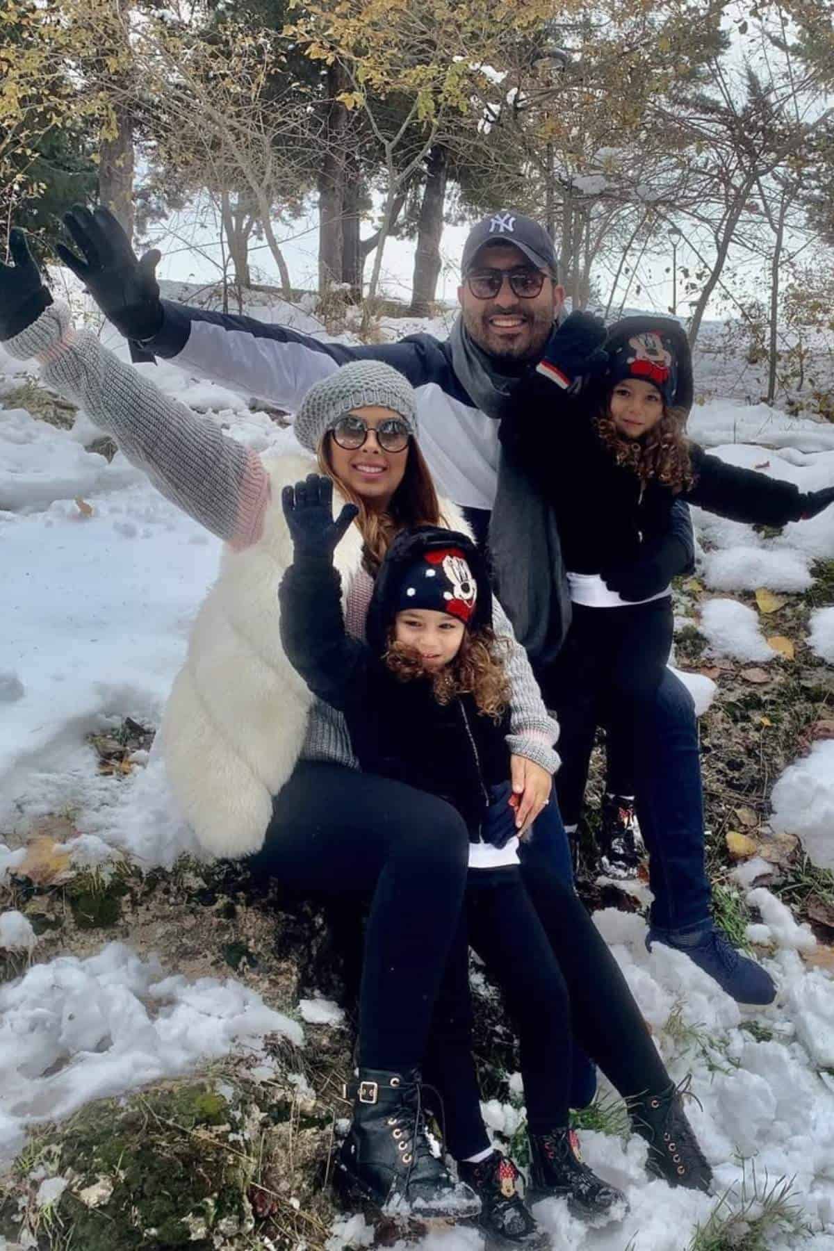 A family of four dressed in winter clothes sits on snowy ground outdoors, smiling and waving at the camera. Trees with some autumn leaves are visible in the background.