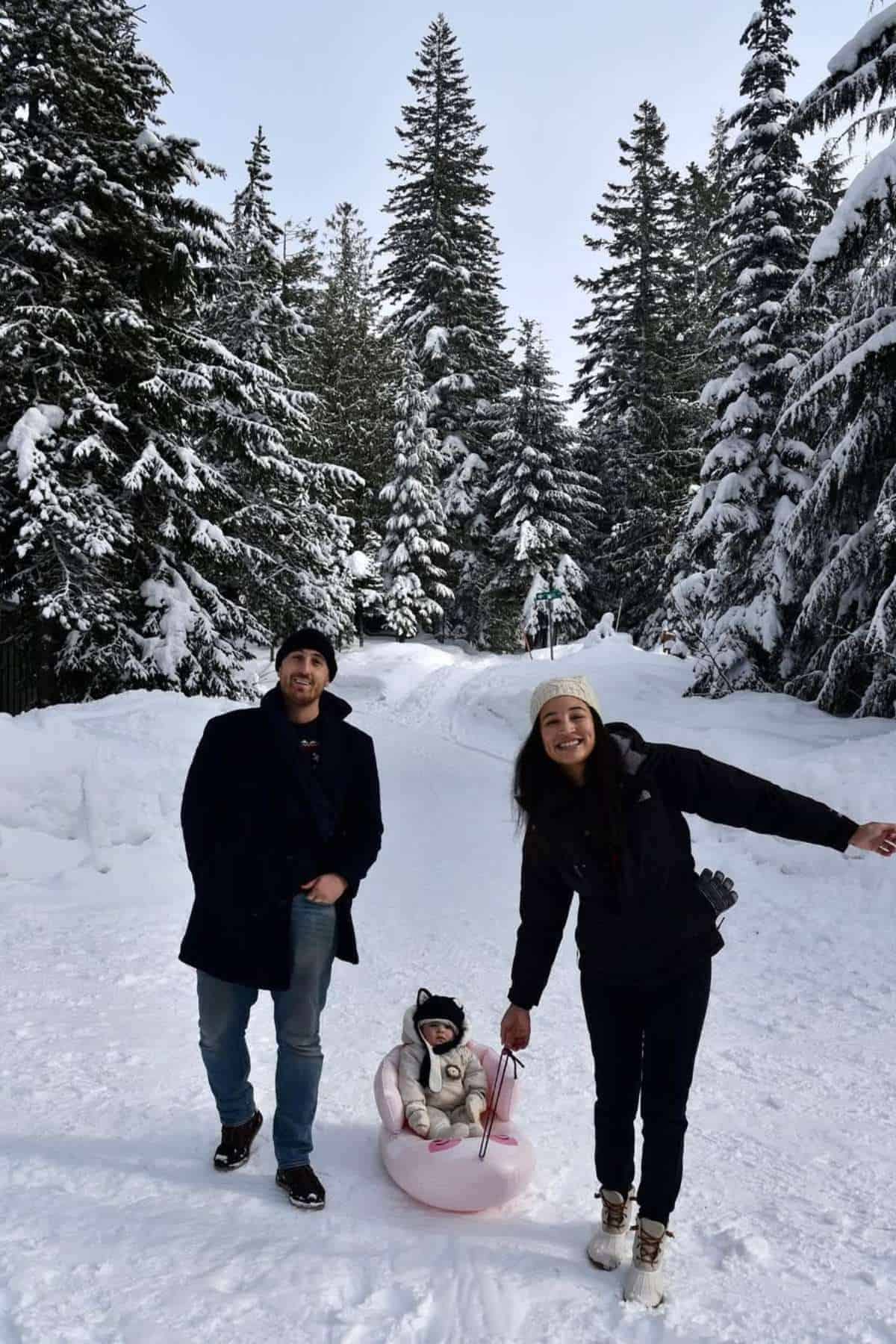 Two adults stand in a snowy forest while a baby in winter clothes sits on a sled. Tall snow-covered trees surround them under a clear sky.