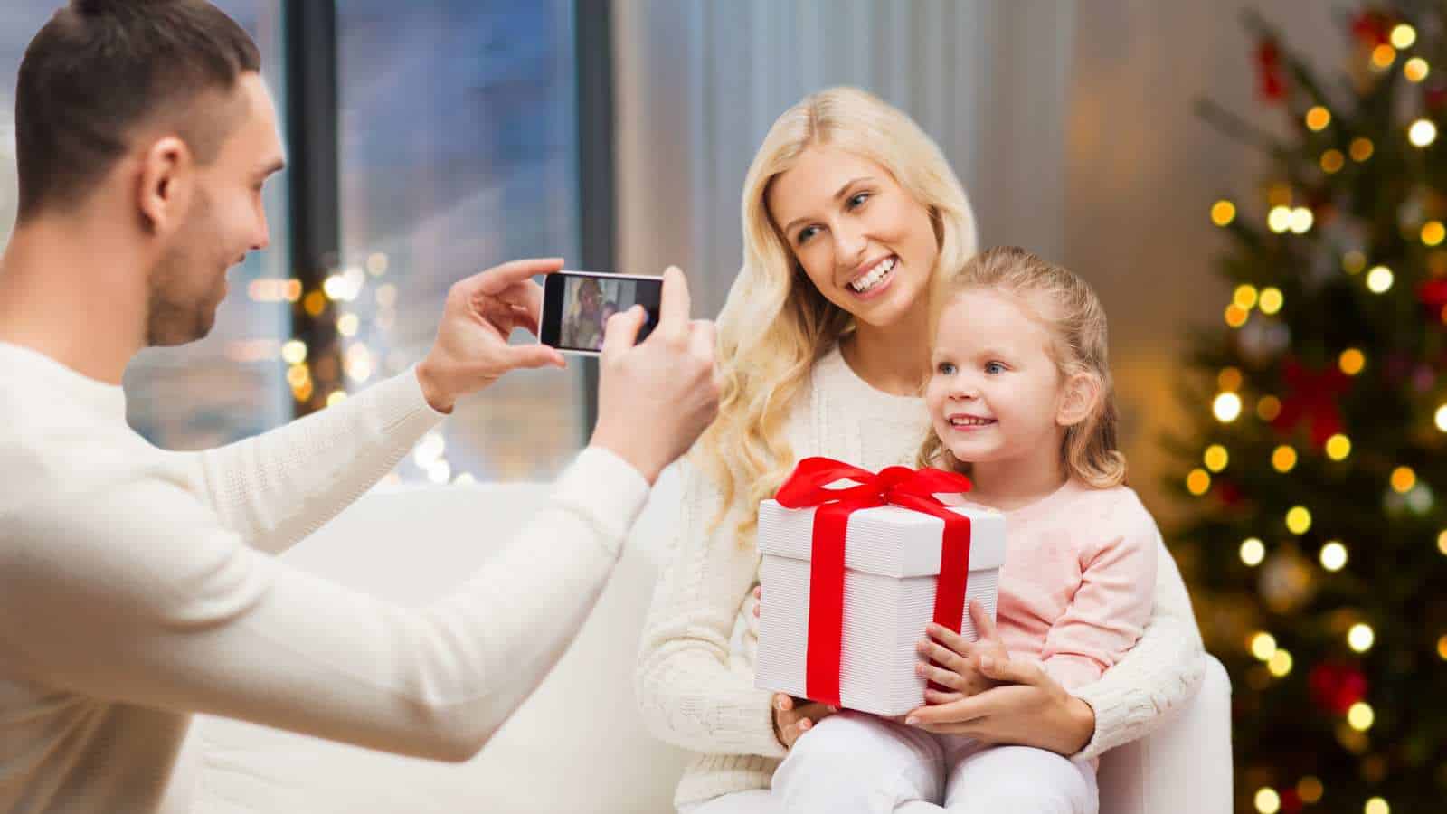 A man takes a photo of a woman and child on a sofa; the child holds a gift box with a red ribbon. A decorated Christmas tree is visible in the background.