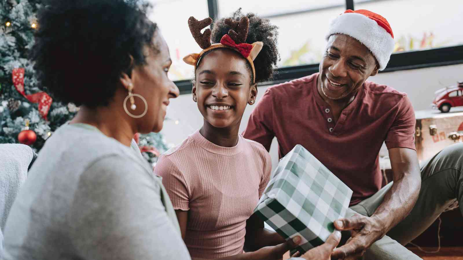 A smiling family sits by a Christmas tree. A child wearing reindeer antlers receives a wrapped present from an adult in a Santa hat while another adult looks on.