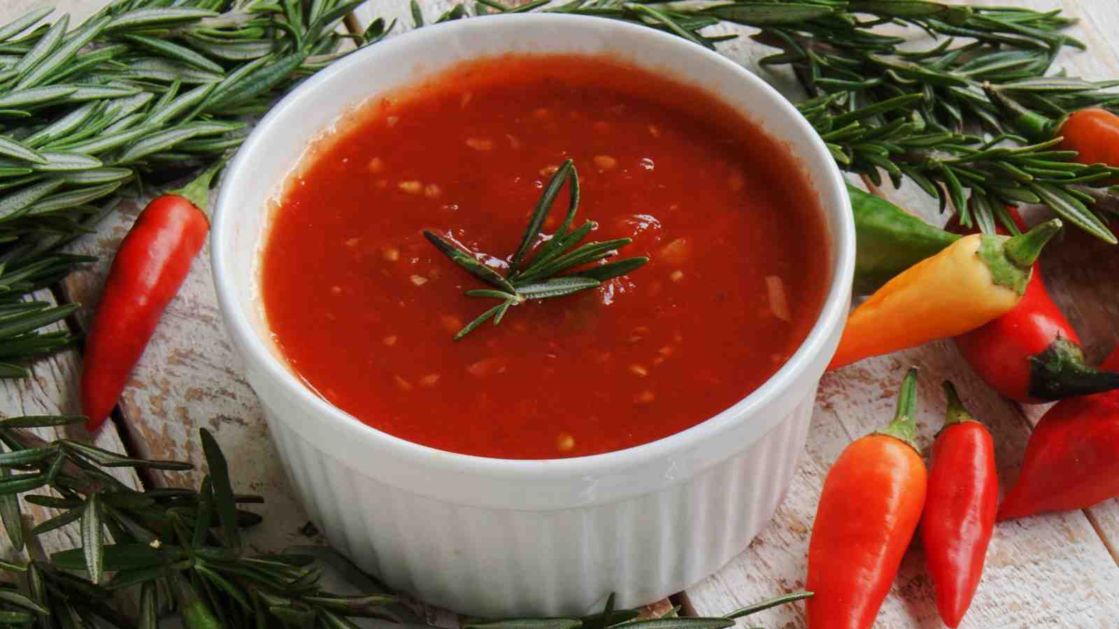 A white ramekin filled with tomato-based sauce, garnished with a sprig of rosemary, surrounded by chili peppers and rosemary sprigs on a wooden surface.